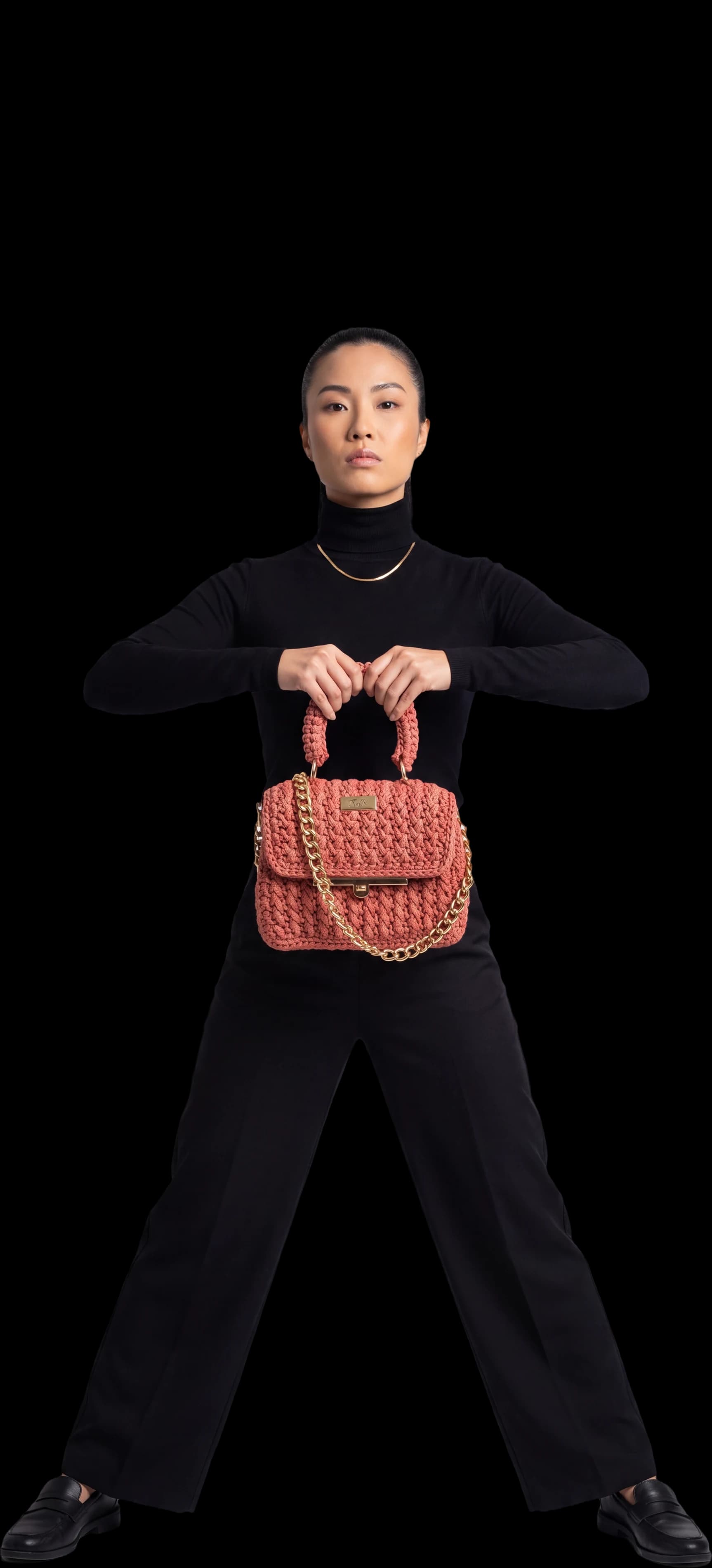 Mulher de preto, de frente, segurando a bolsa Singapura coral na altura do peito, imagem ilustrativa