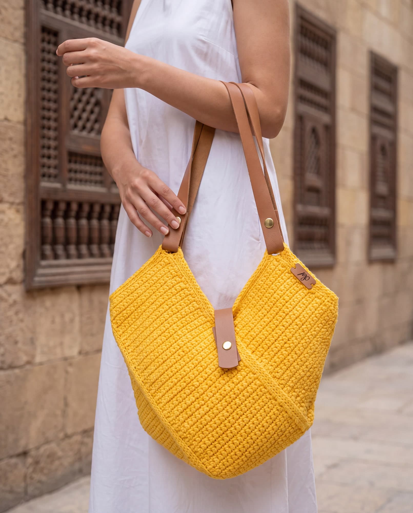 Mulher de vestido branco com a bolsa Cairo amarela no antebraço, numa viela de pedra com treliças de madeira, imagem ilustrativa