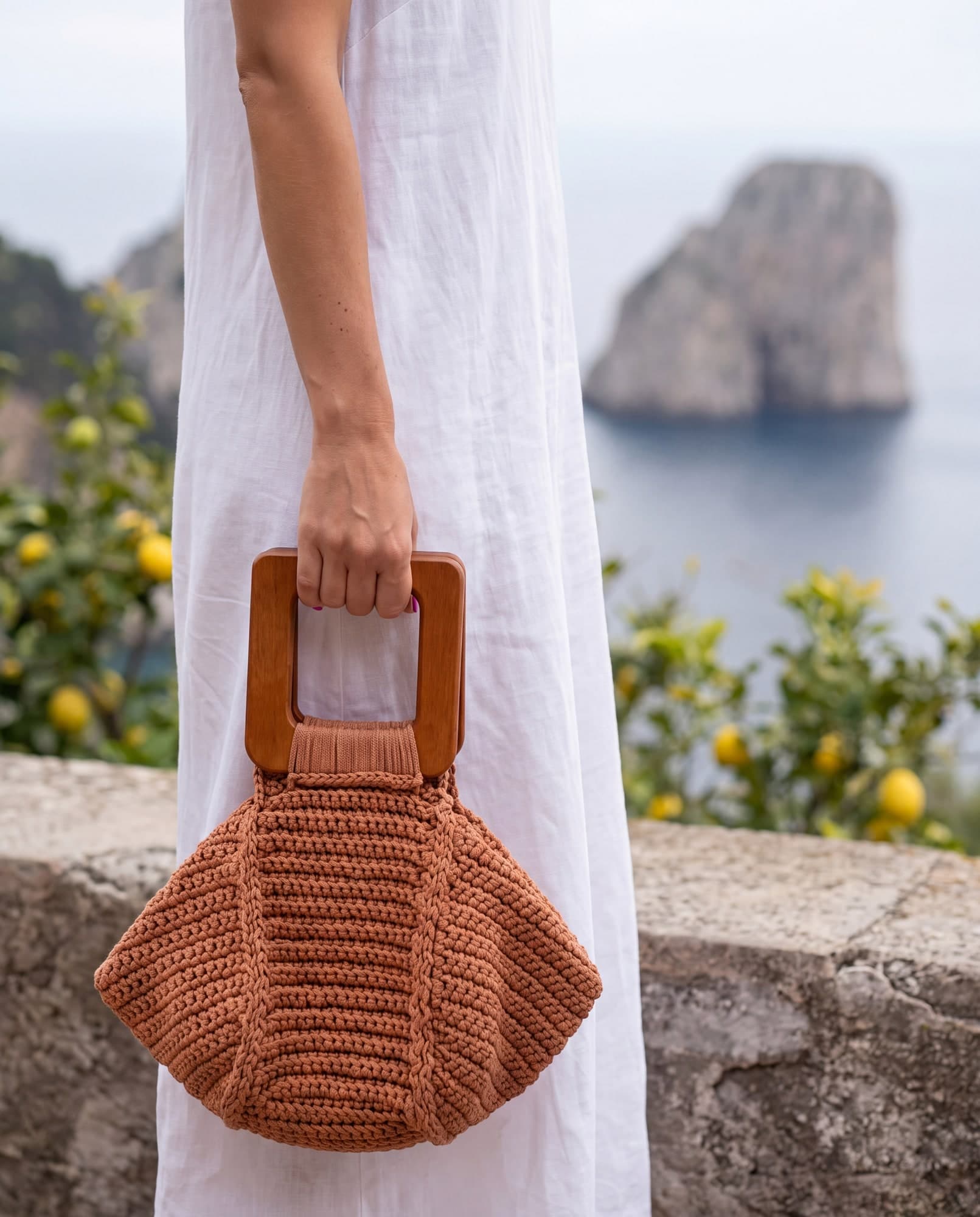 Mulher de vestido de linho branco segurando a bolsa Capri terracota pela alça de madeira, num terraço com limoeiros e o mar ao fundo, imagem ilustrativa