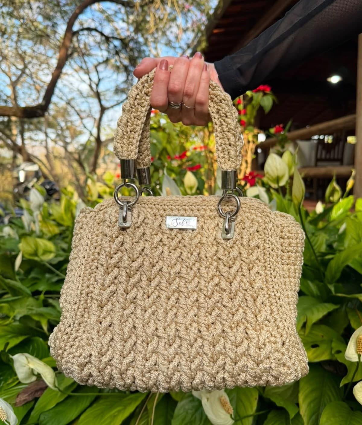 Bolsa Dublin bege em ponto trança, segurada pela alça em um jardim