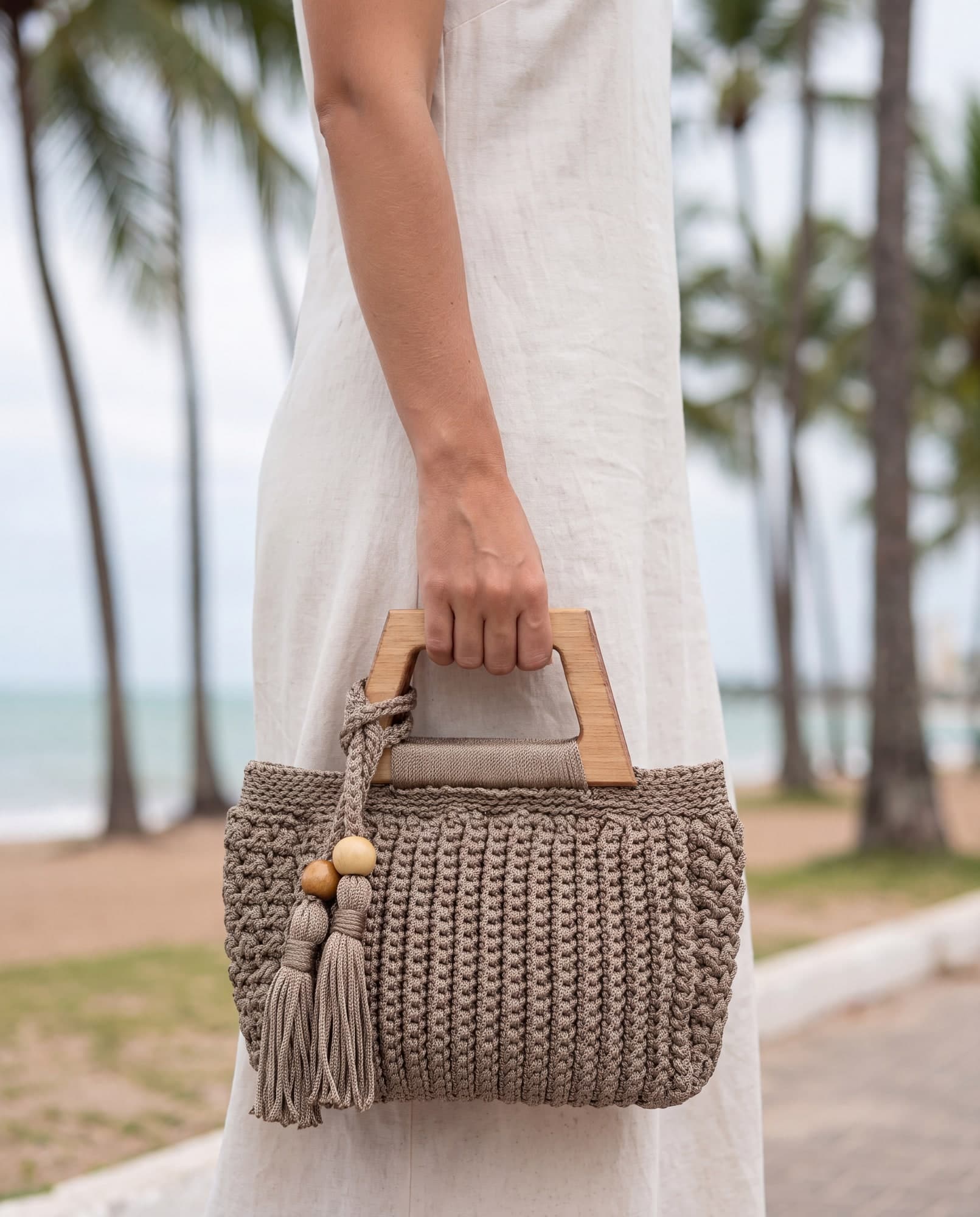 Mulher de vestido off-white segurando a bolsa Maceió fendi pela alça de madeira, num calçadão de coqueiros, imagem ilustrativa