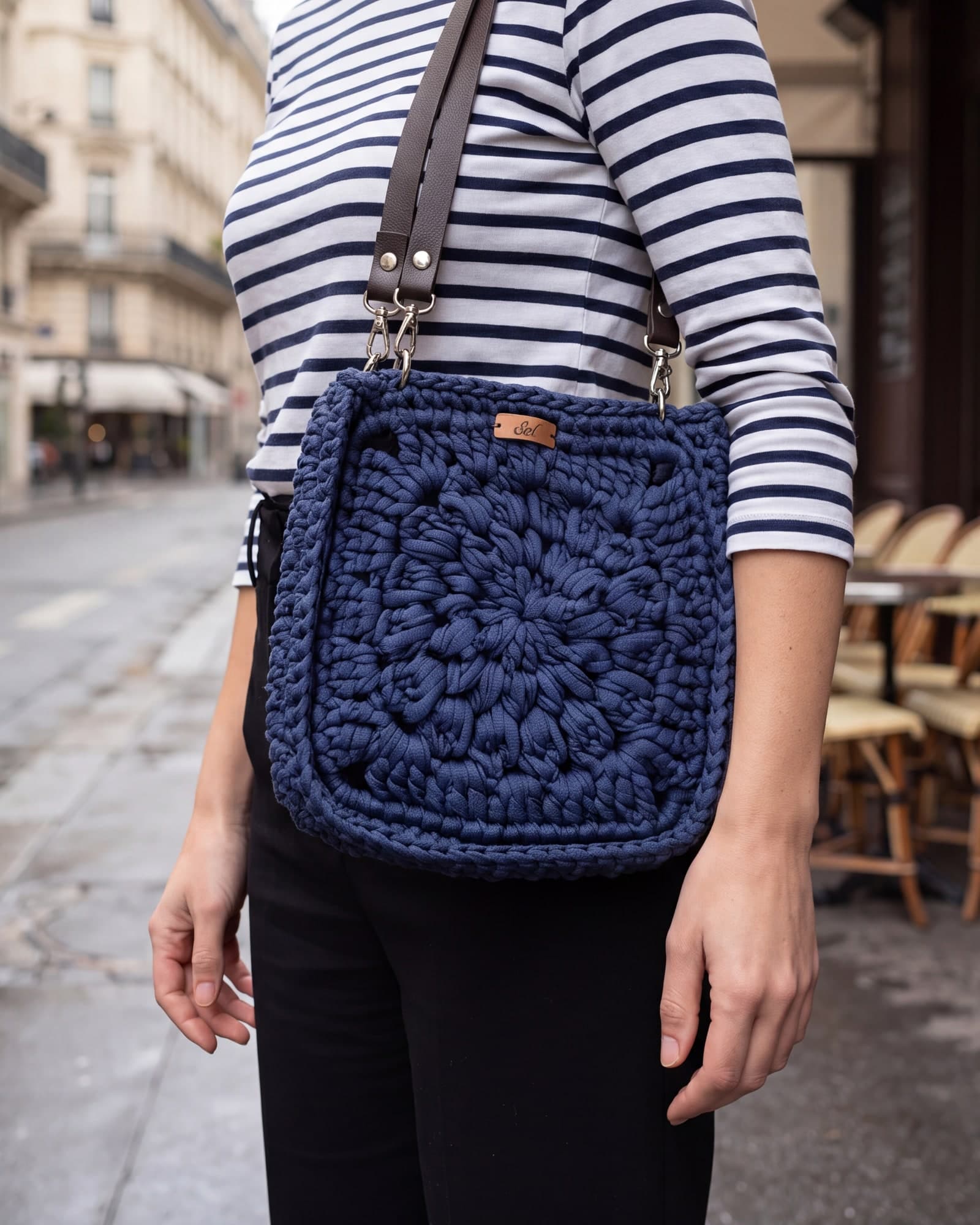 Mulher de blusa listrada com a bolsa Paris azul-marinho no ombro, numa rua de prédios haussmannianos, imagem ilustrativa